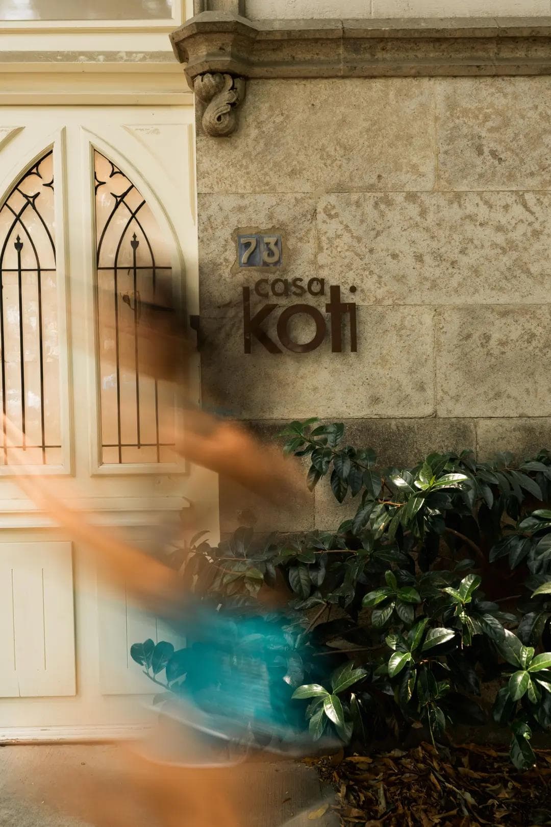 Facade of Casa Koti, a serene wellness space with natural stone walls and greenery, capturing a sense of calm and balance as a blurred figure passes by, symbolizing movement and vitality.